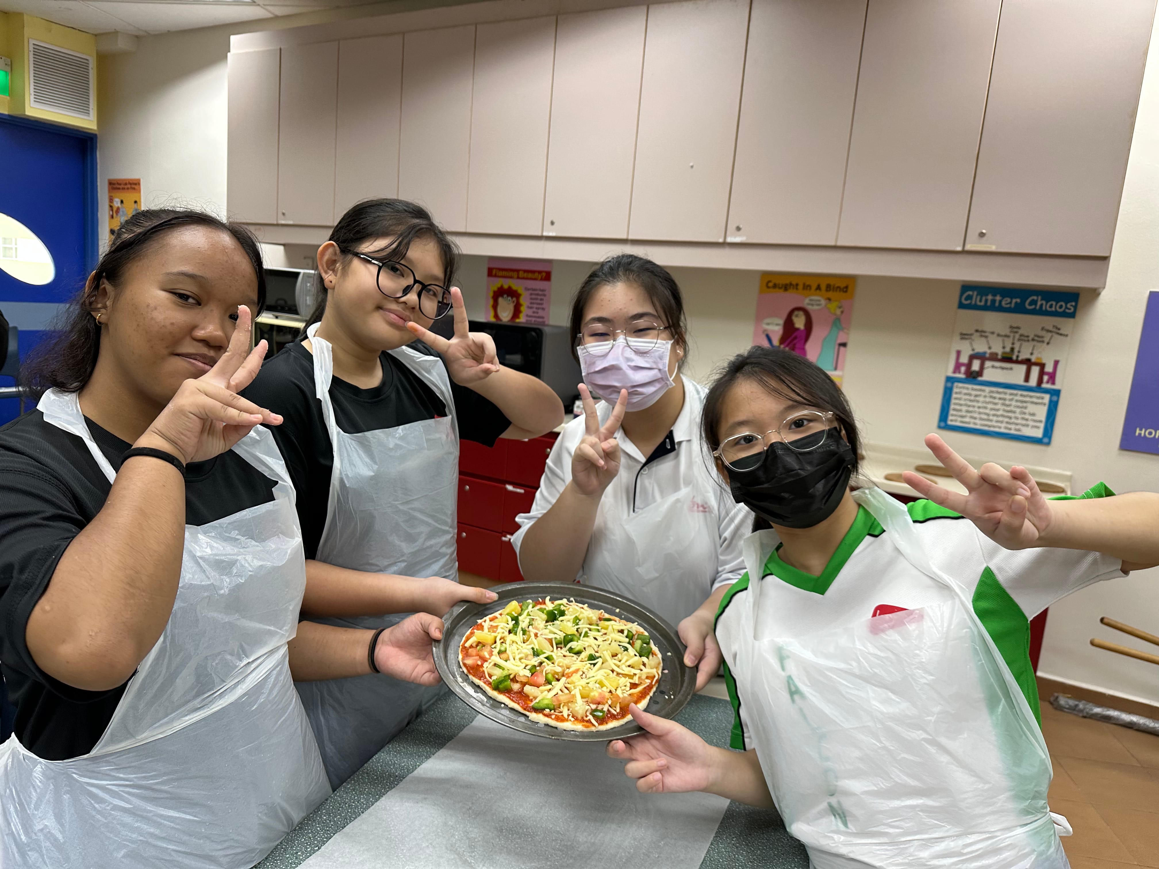 Four teenage girls in aprons hold an uncooked homemade pizza while posing with peace signs in a kitchen classroom.